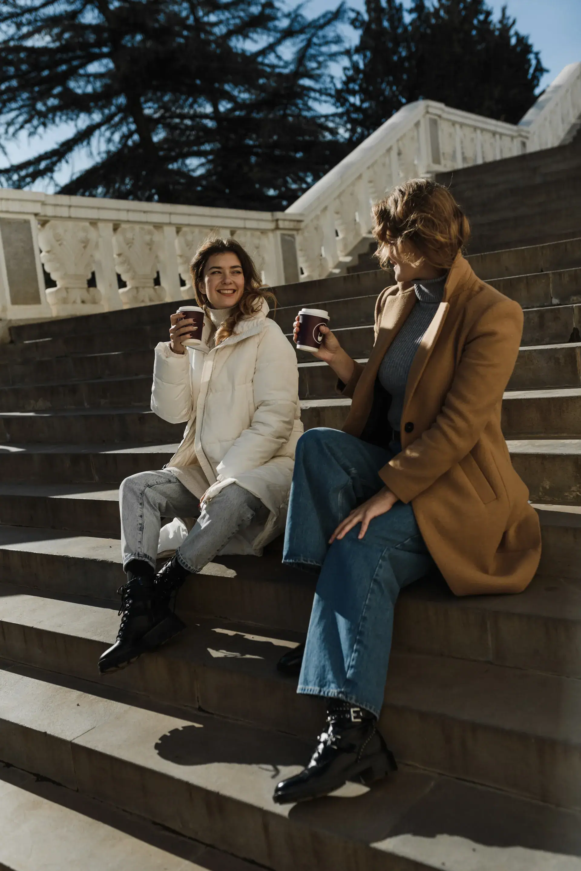 Dos estudiantes riéndose con un café en unas escaleras de piedra un día de sol